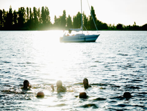 Mensen zwemmen in een meer bij zonsondergang; een zeilboot vaart op de achtergrond.