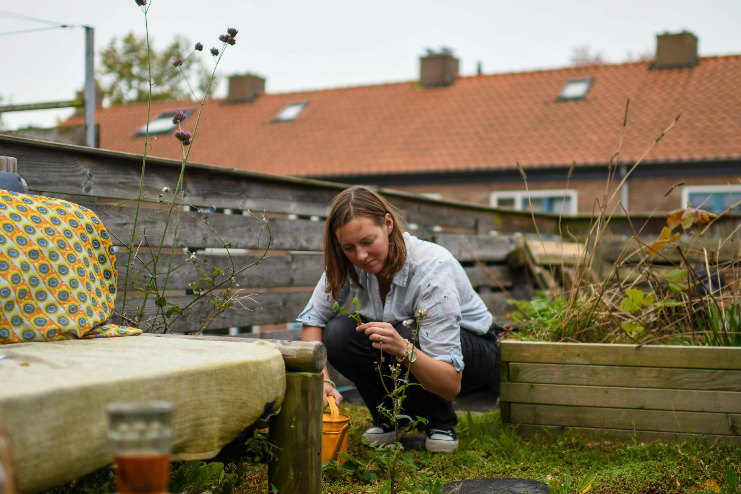 Vrouw verzorgt planten in een tuin met houten schutting en bakstenen huis op de achtergrond.