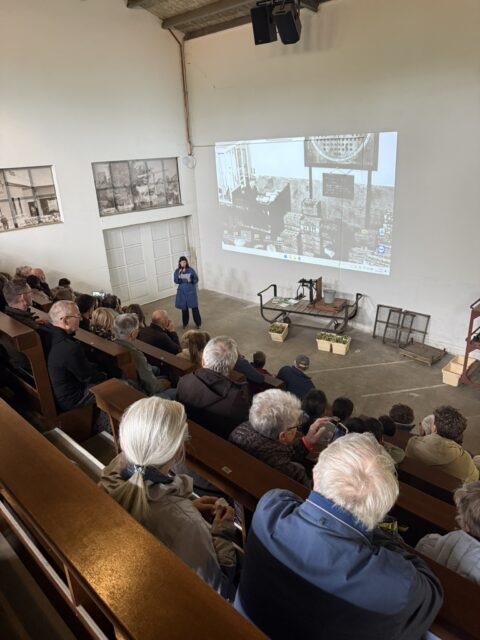 Een spreker presenteert voor een publiek in een auditorium; een zwart-wit afbeelding geprojecteerd.