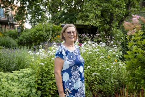 Vrouw in blauwe jurk glimlacht in groene tuin met bloeiende bloemen.