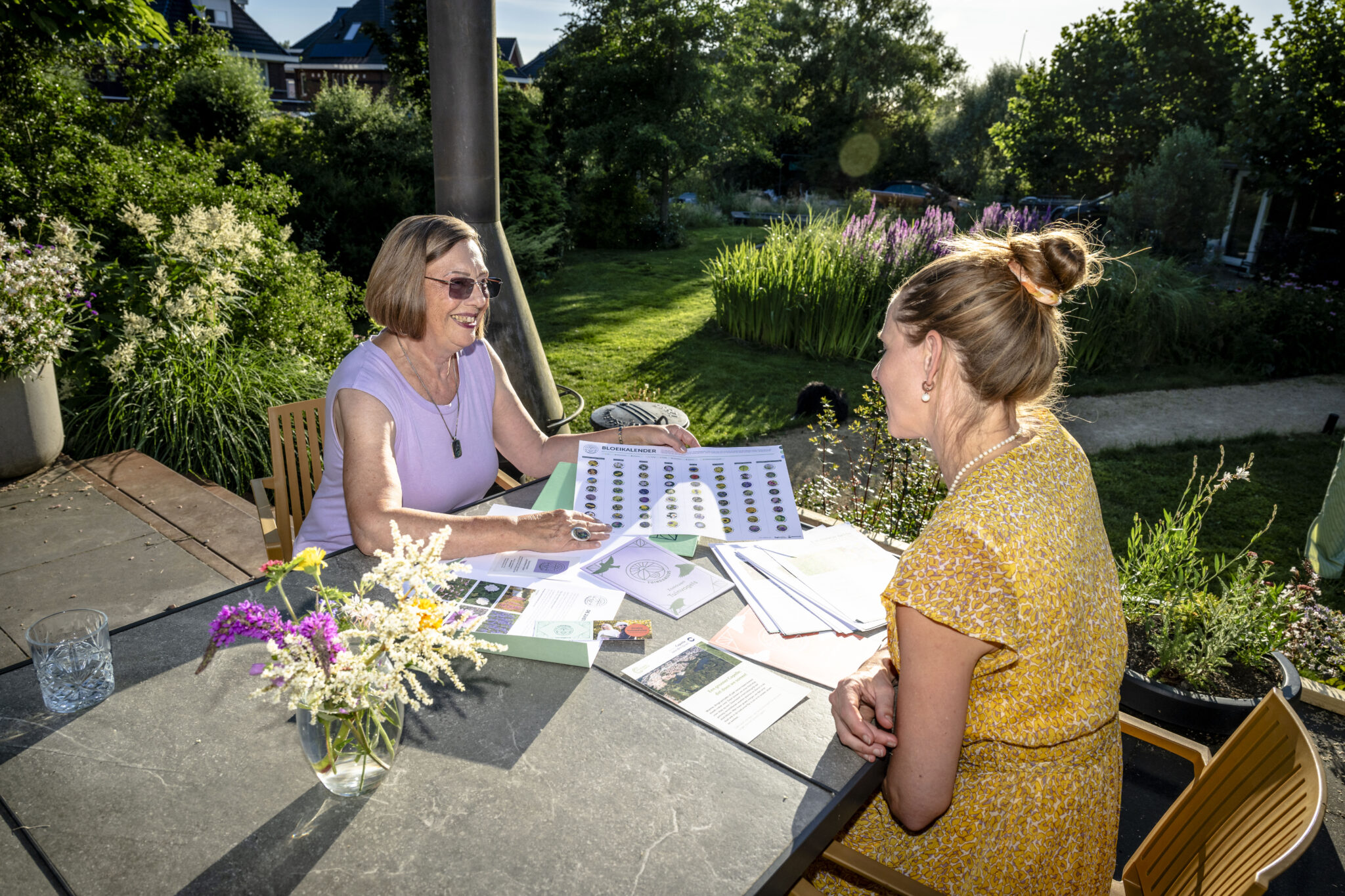 Twee vrouwen bespreken diagrammen aan een buitentafel in een tuin, omgeven door groen.