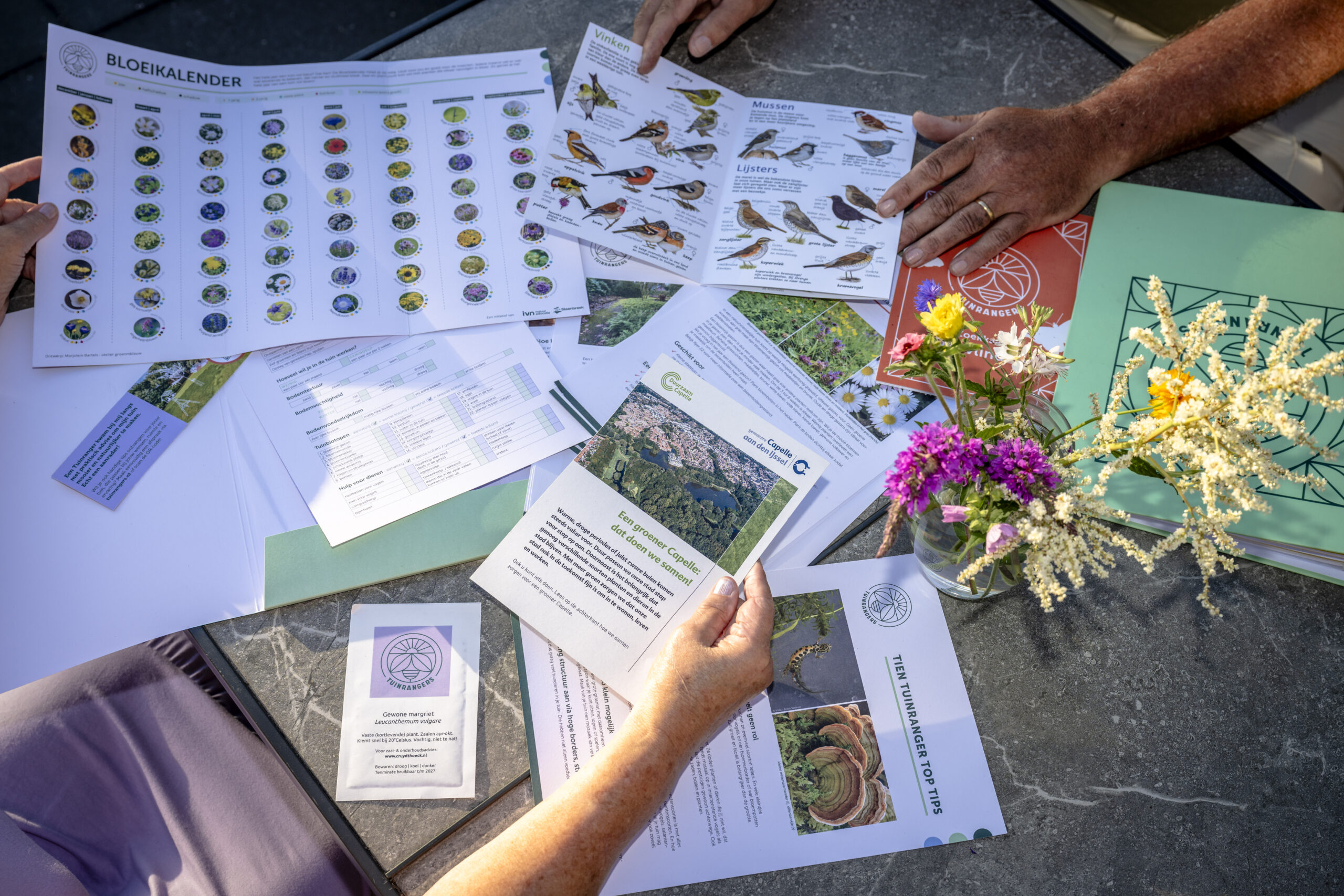 Papieren en bloemen op een tafel, met bloeikalender en vogelgids in gebruik.