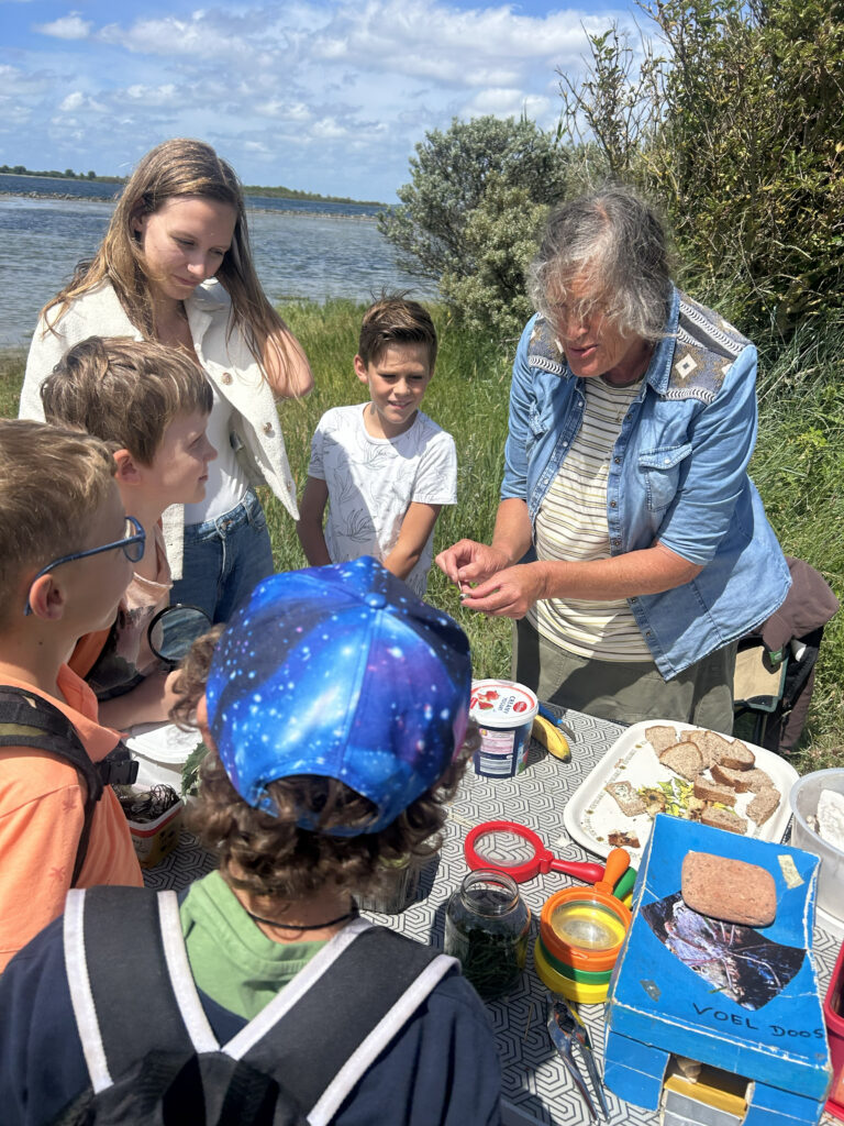Groep kinderen en een volwassene verzamelen zich rond een tafel met natuurbenodigdheden aan het water.
