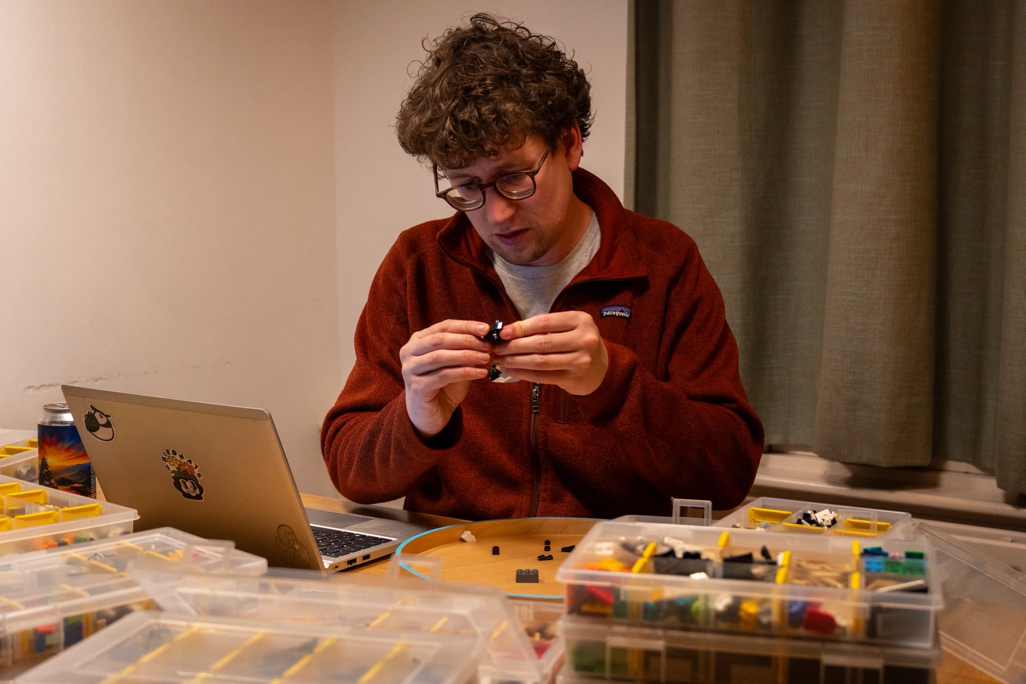 Persoon bouwt met LEGO-steentjes aan tafel, laptop en opbergdozen op achtergrond.