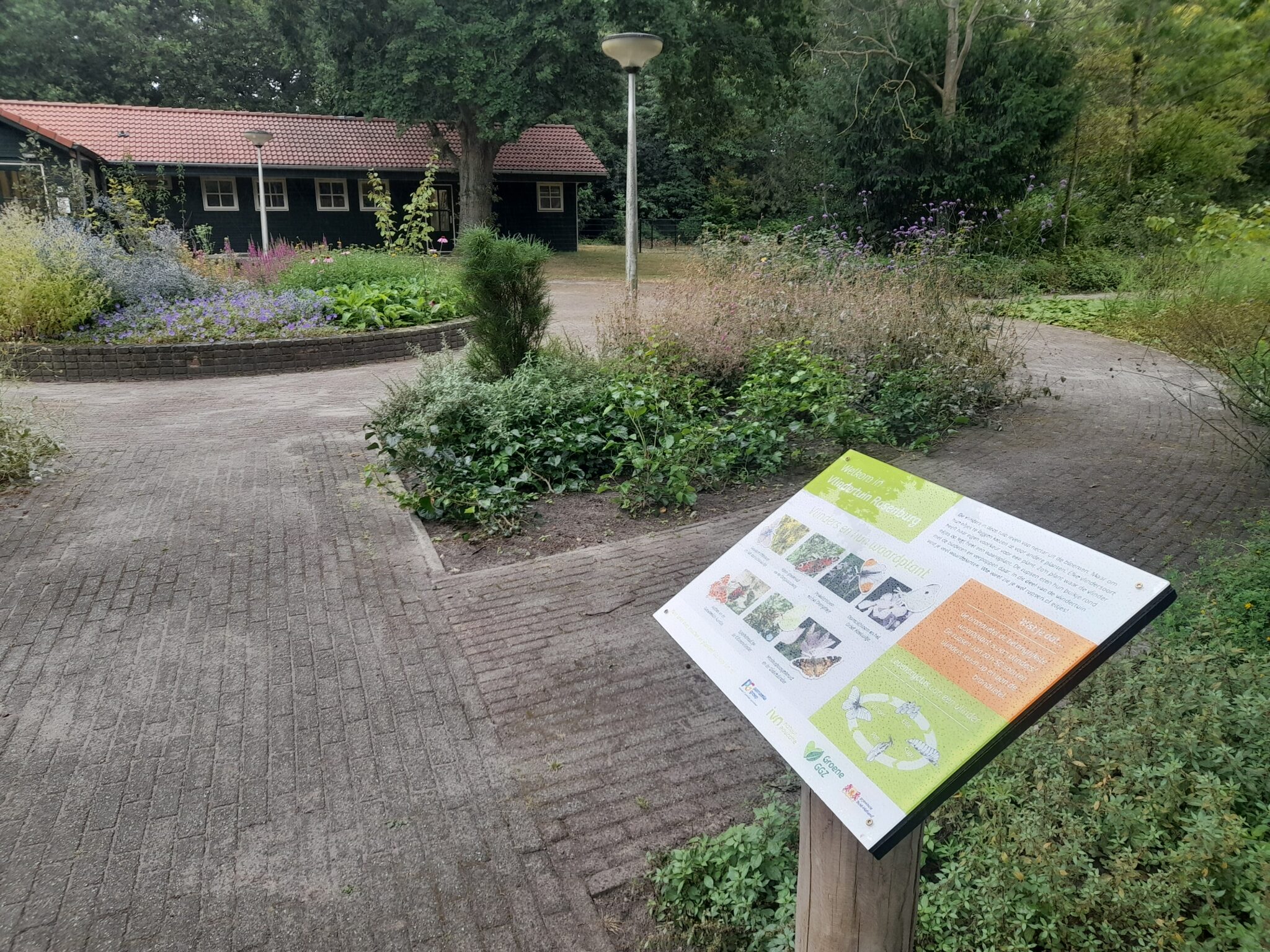 Informatiebord bij een tuinpad met bloemperken, tegenover een gebouw met een rood dak.