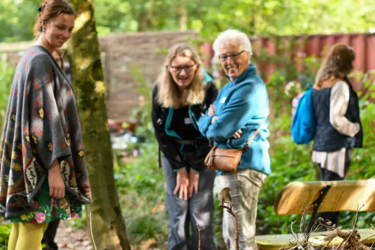 Groepje mensen lacht samen in een groene tuin naast een bankje.