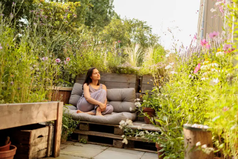 Vrouw ontspant op een bank in een weelderige tuin met veel groene en bloeiende planten.