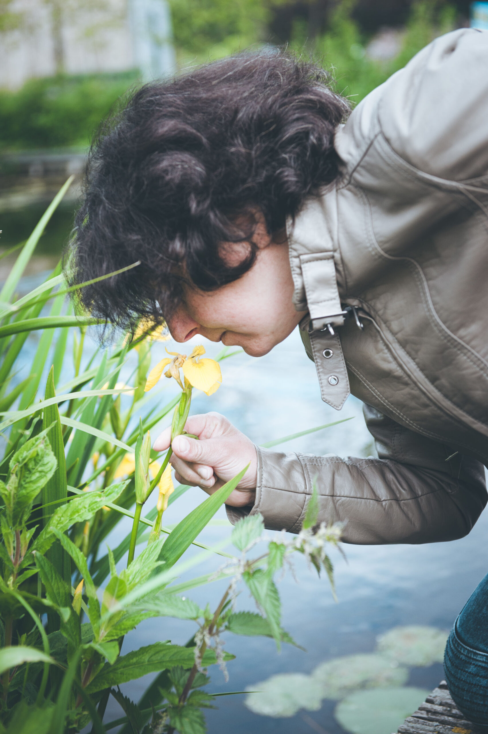 Persoon ruikt aan een gele bloem bij het water, gekleed in een jas.