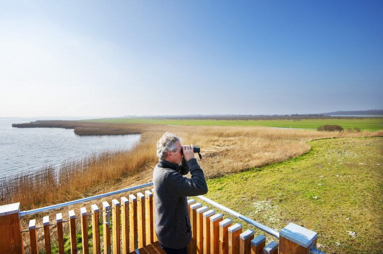 Man met verrekijker kijkt uit over grasland en water vanaf houten platform.