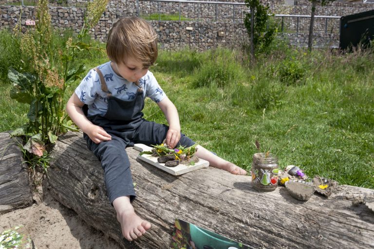 Kind zit op boomstam, speelt met bloemen en bladeren op een bordje in een tuin.