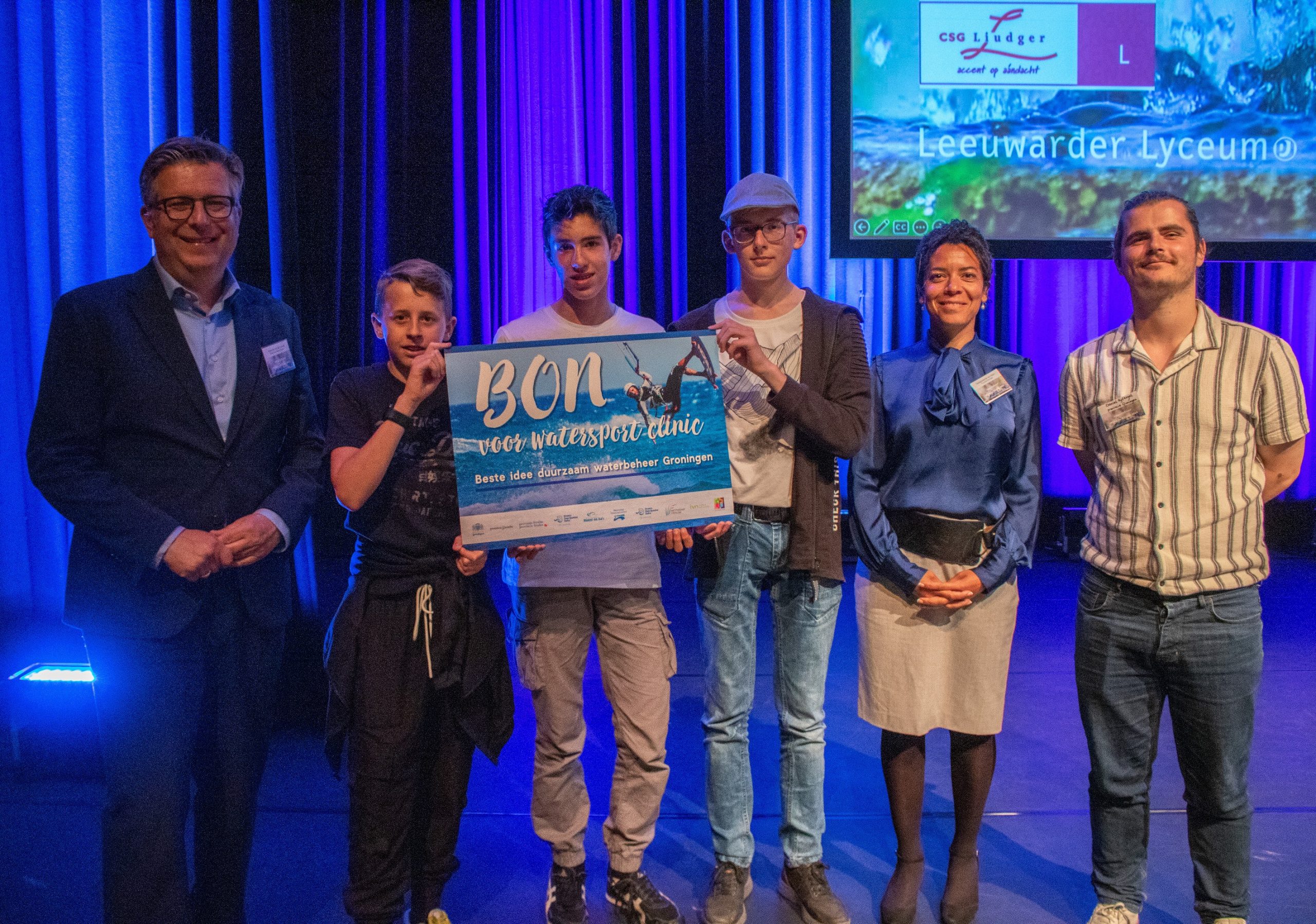 Een groep mensen poseert op een podium met een poster voor een watersportclinic.