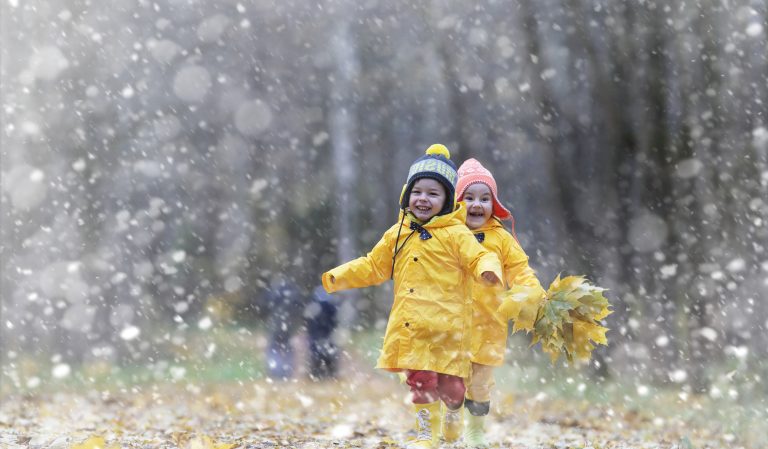 Kinderen in gele regenjassen rennen lachend tijdens lichte sneeuwval, omringd door herfstbladeren.