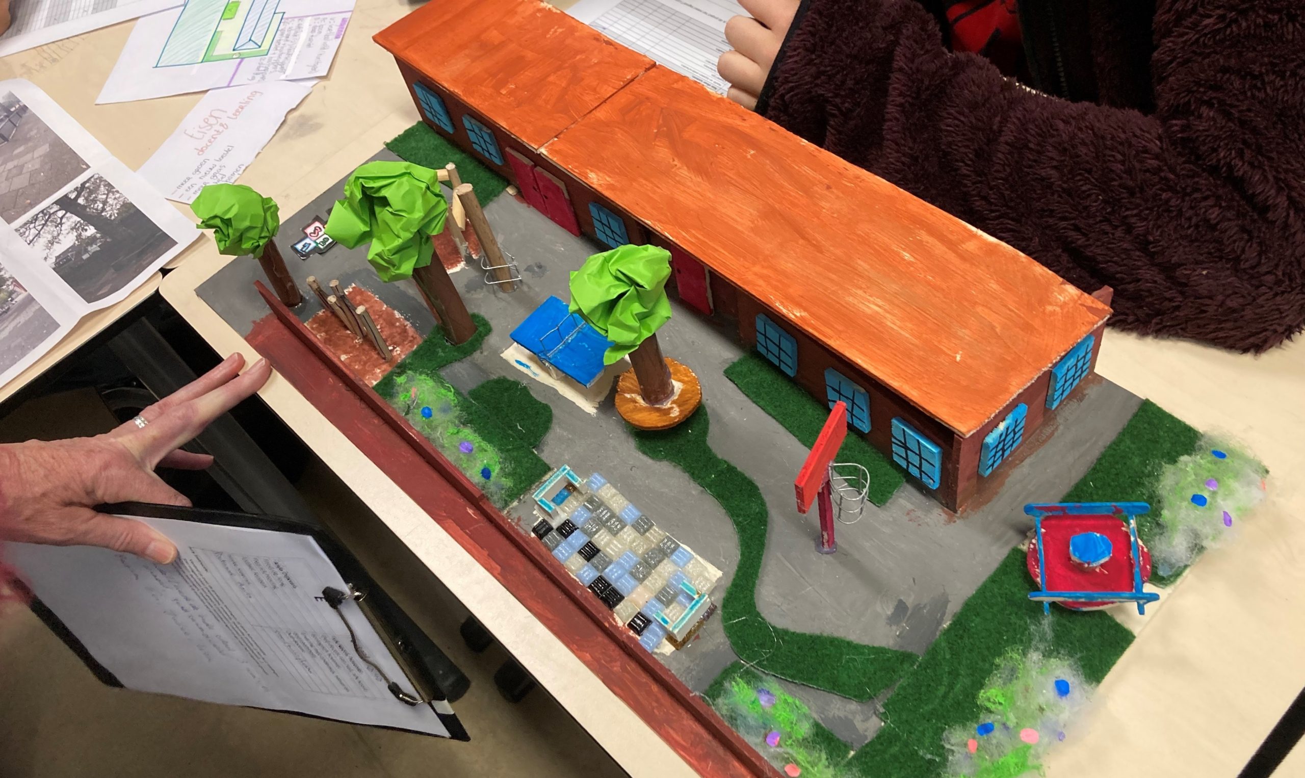 Miniatuurmodel van een school met oranje dak, bomen en speelelementen. Persoon met clipboard erbij.