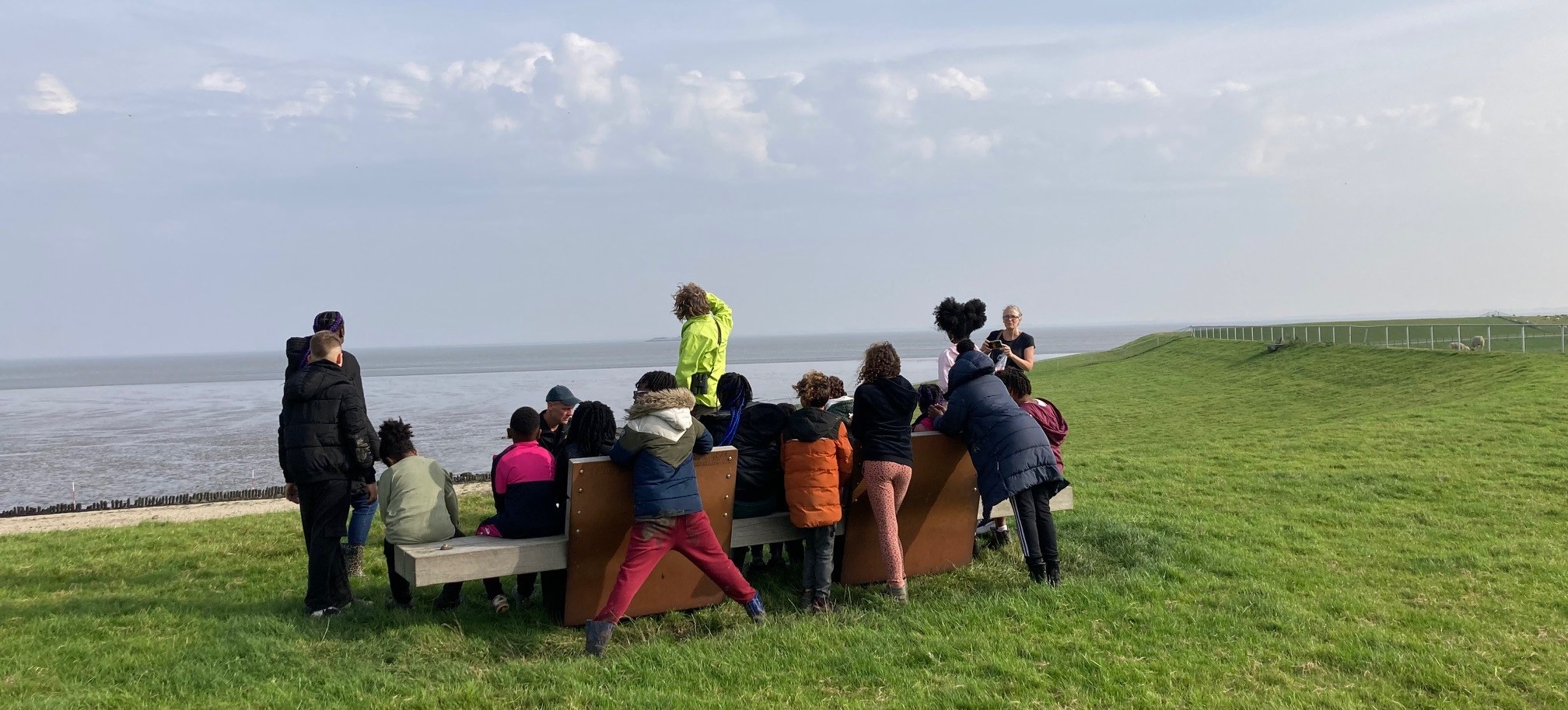 Groep kinderen op een bankje kijkt uit over een grasheuvel en zee, onder bewolkte lucht.