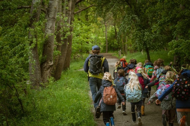 Groep kinderen met rugzakken loopt met een volwassene op een bospad omringd door groen.