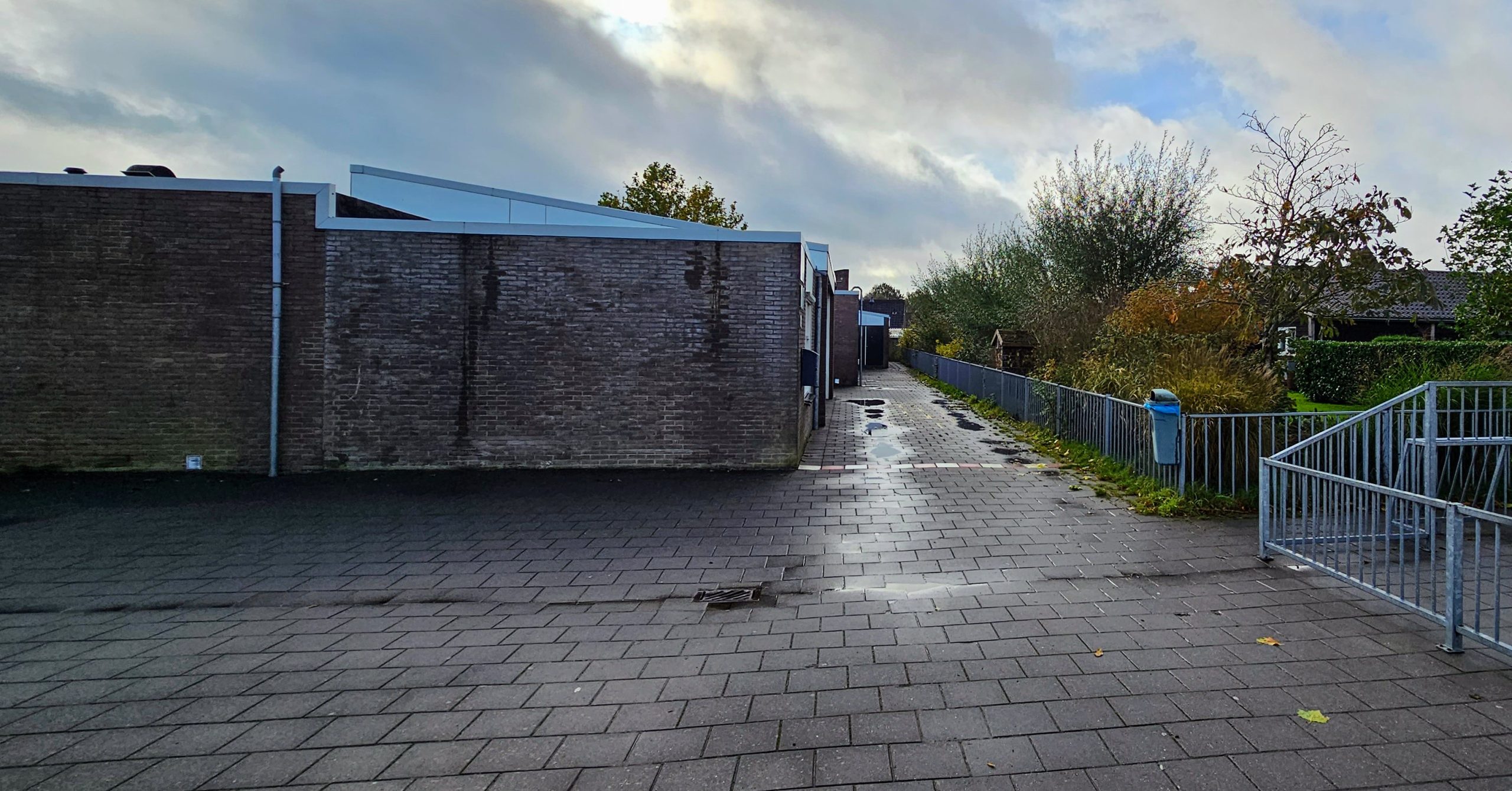 Verlaten, natte bestrating naast een grauwe bakstenen muur en hek met bomen op de achtergrond.