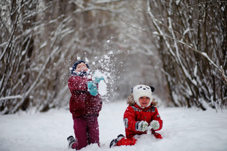 Twee kinderen spelen in de sneeuw, gekleed in warme kleding op een besneeuwd bospad.