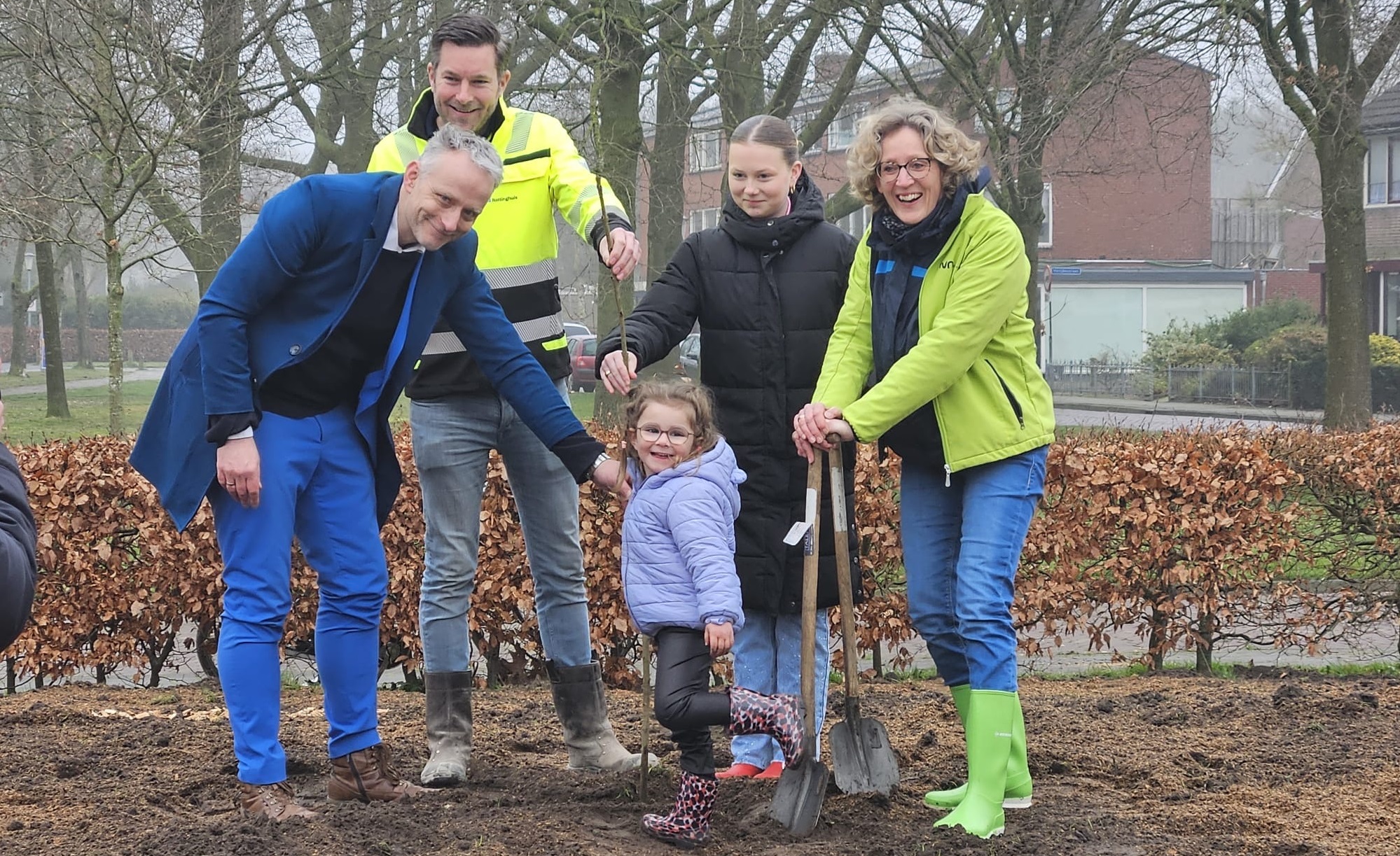 Mensen planten samen een jonge boom in een park, omringd door bomen en huizen.