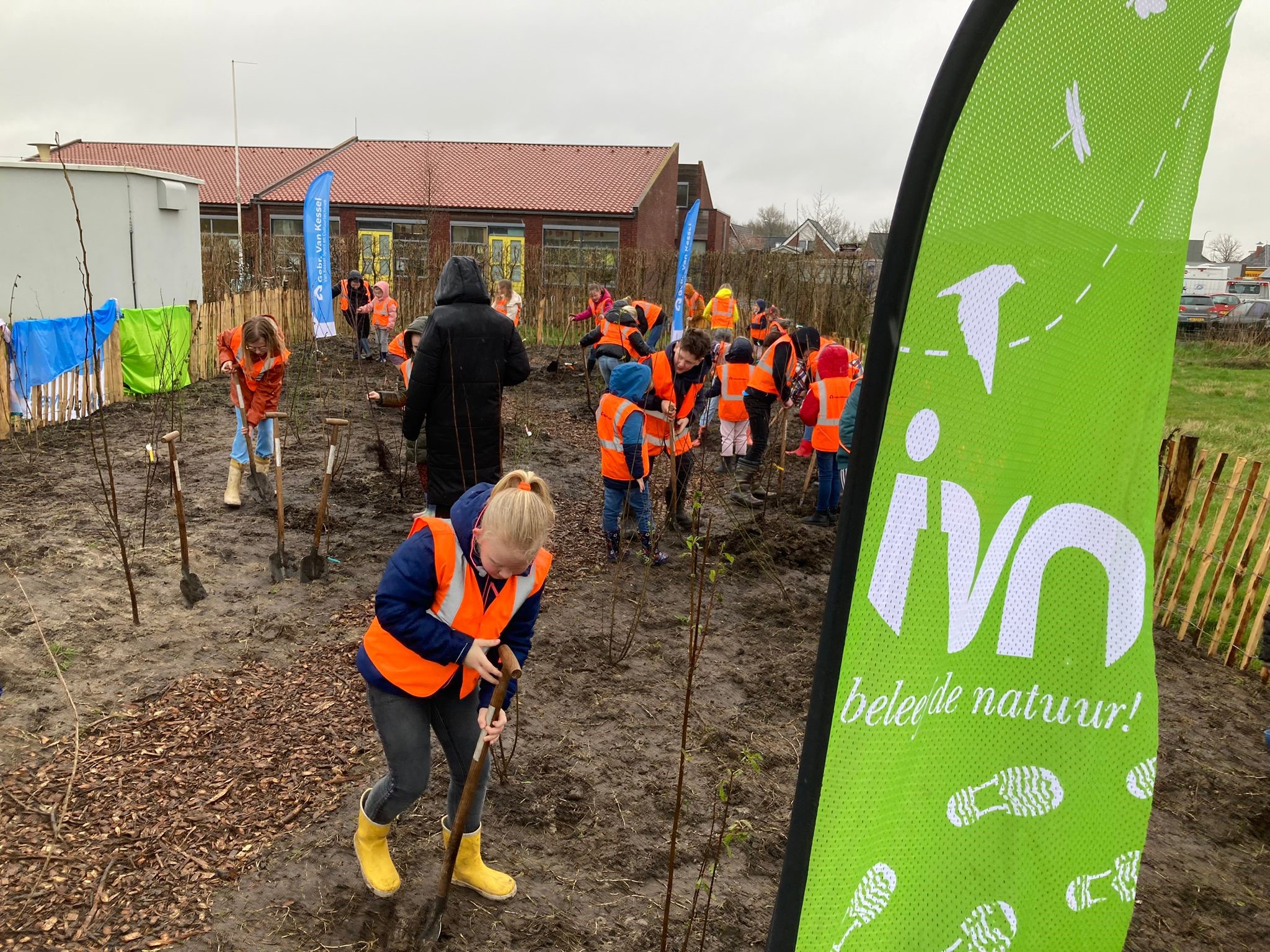 Kinderen planten bomen in een modderige tuin, in oranje hesjes, naast een groene IVN vlag.