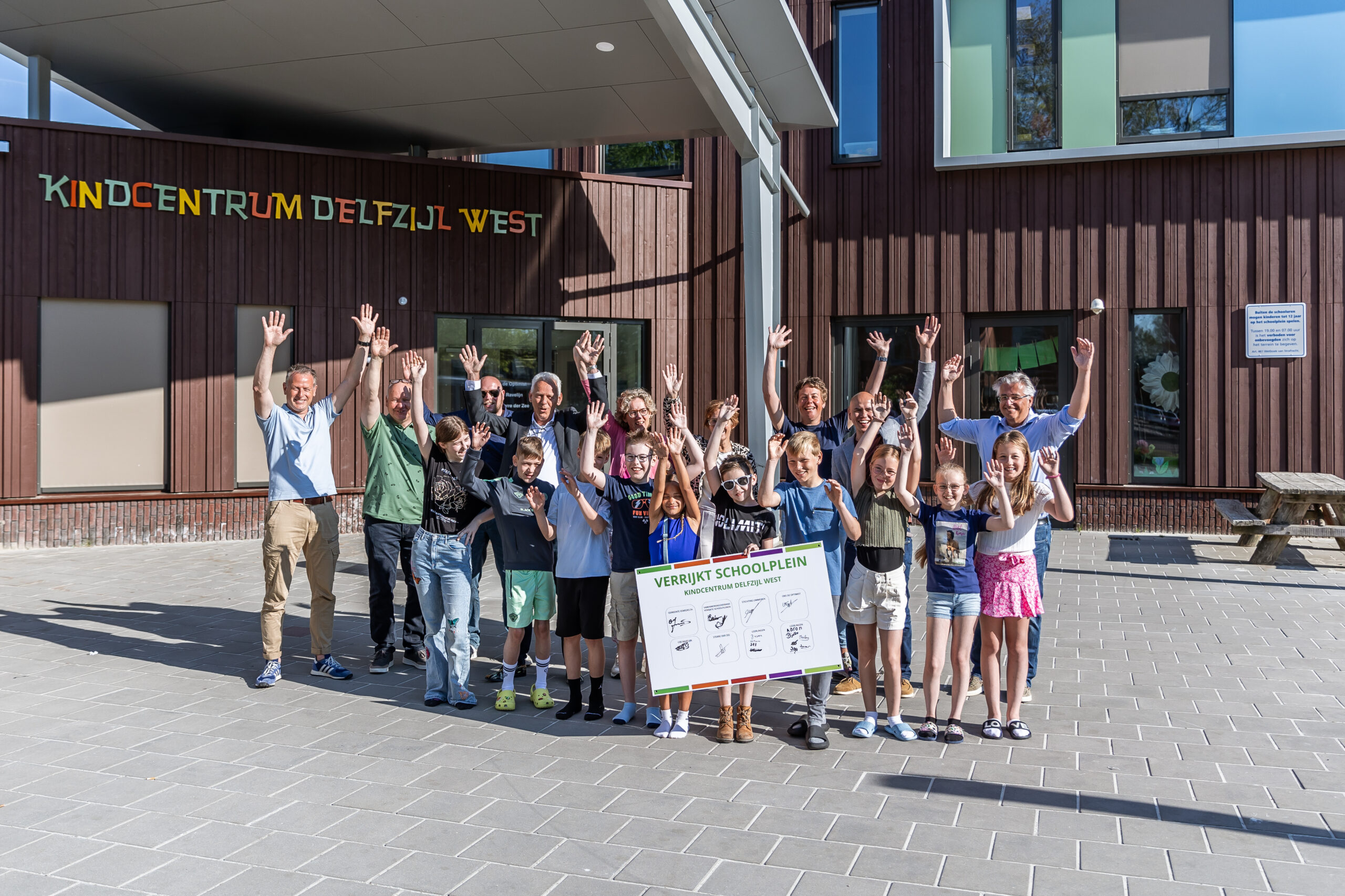 Groep juichende mensen voor Kindcentrum Delfzijl West met een bord voor een verrijkt schoolplein.
