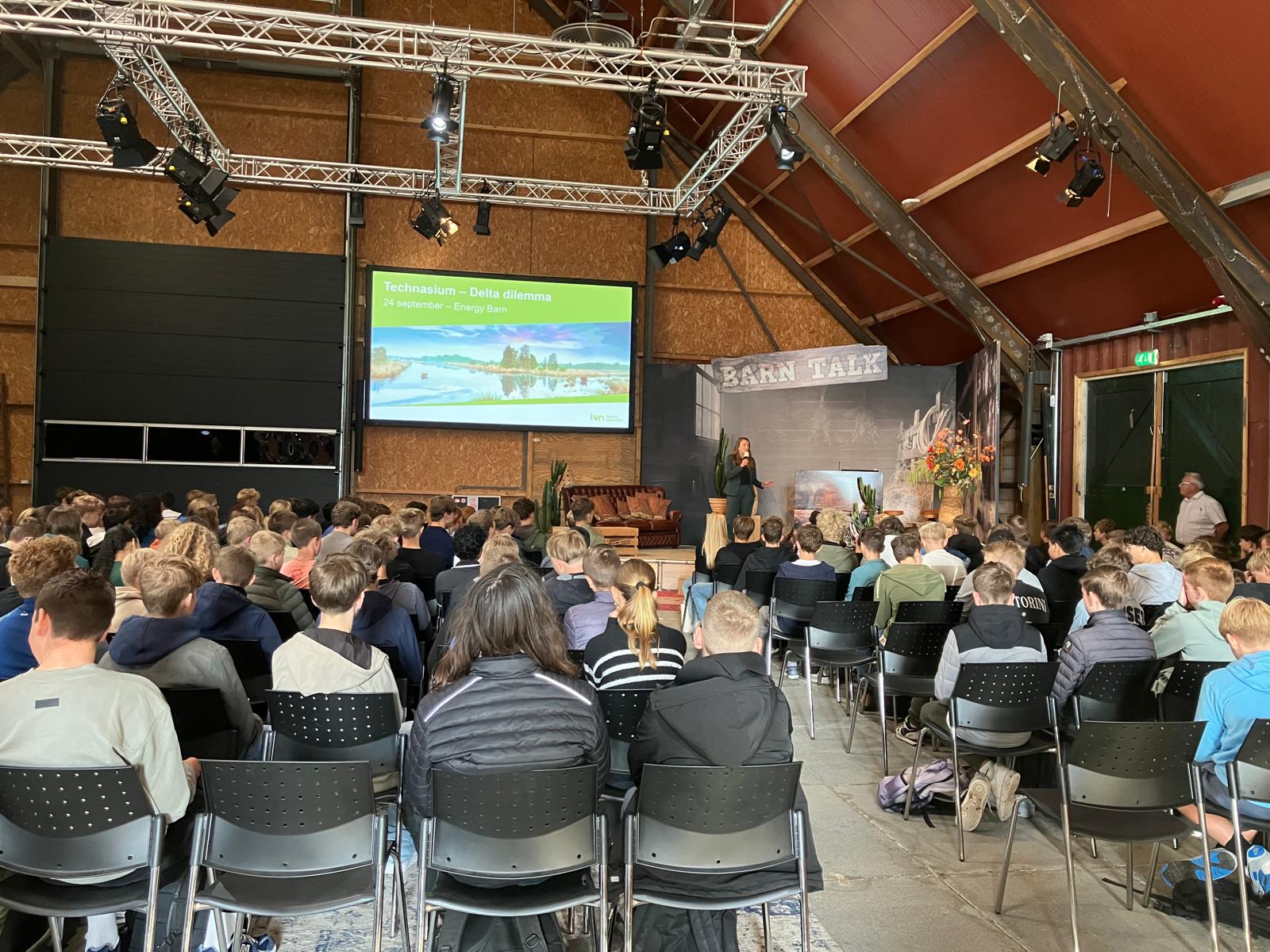 Publiek luistert naar lezing in een schuur met een scherm dat het "Delta dilemma" presenteert.