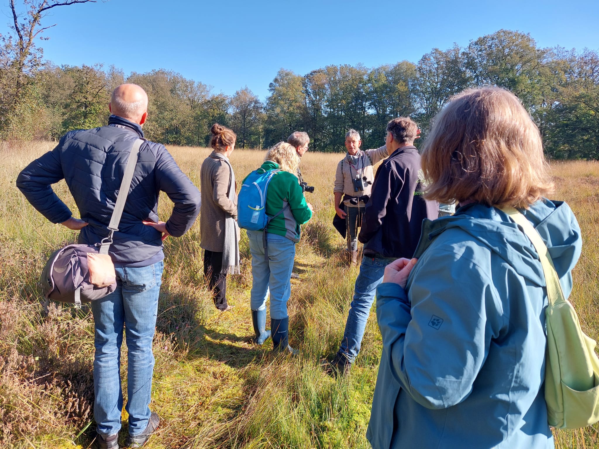 Mensen staan in een open veld en luisteren naar een gids onder een blauwe hemel.