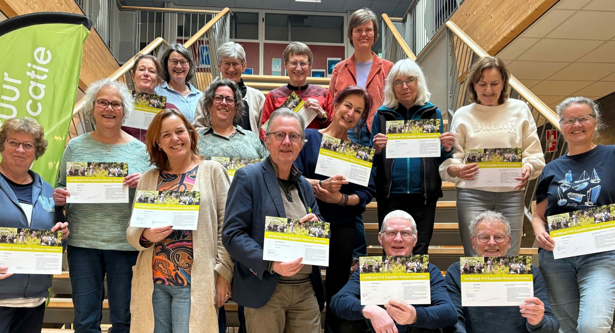 Groep mensen op een trap houdt certificaten vast voor IVN Expeditie Nieuwe Gezichten, met een groene vlag links.