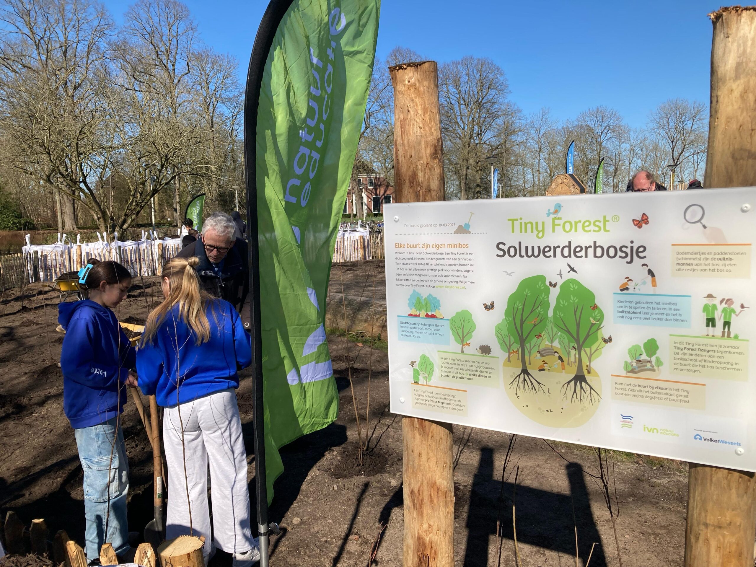 Mensen planten een boom in Tiny Forest Solwerderbosje, met een informatiebord en banner op de achtergrond.