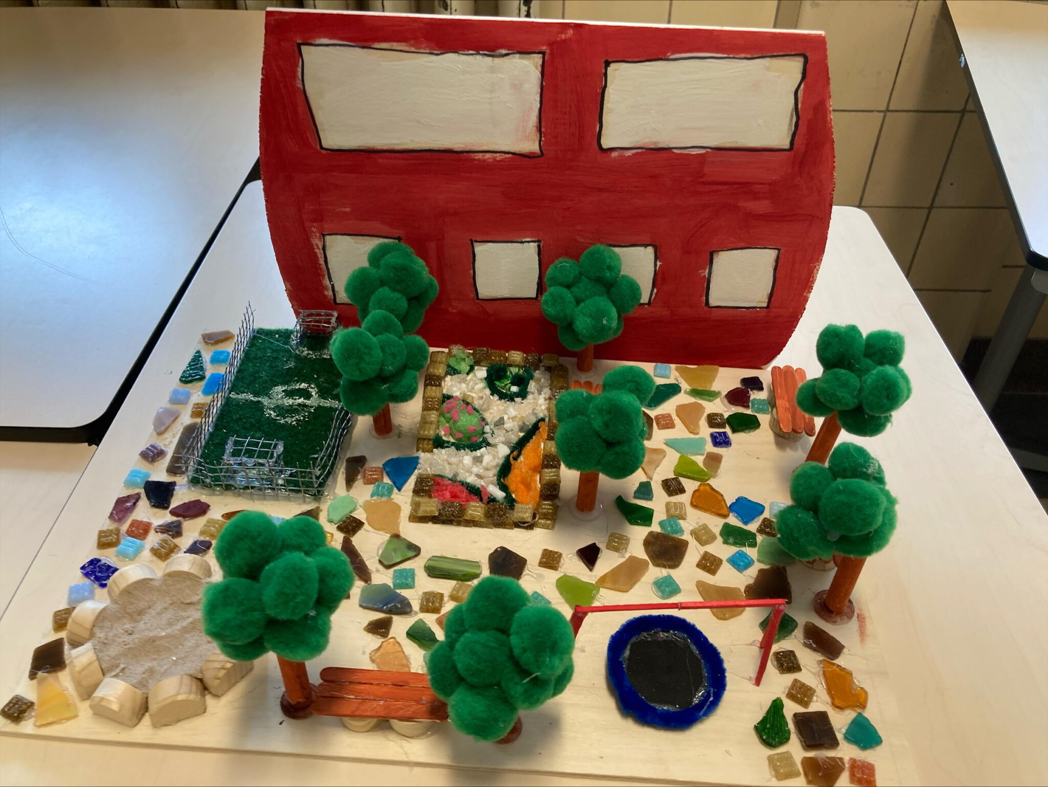 Maquette van schoolplein met rode school, bomen van groene pompons, speeltuin en gekleurde tegels.