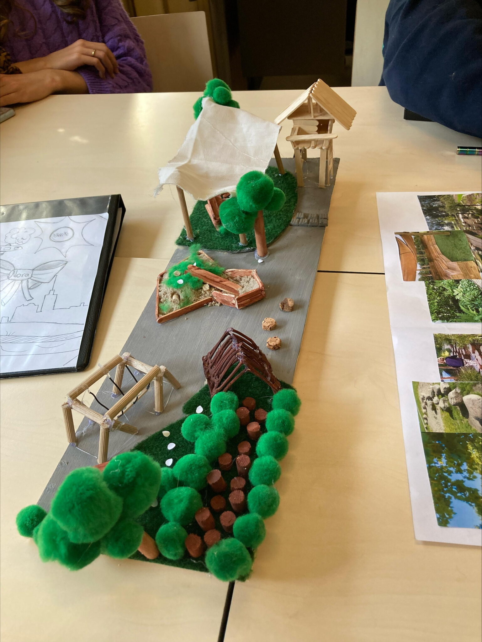 Een maquette van een landschap met bomen, tent en gebouw, omringd door mensen die het bekijken.