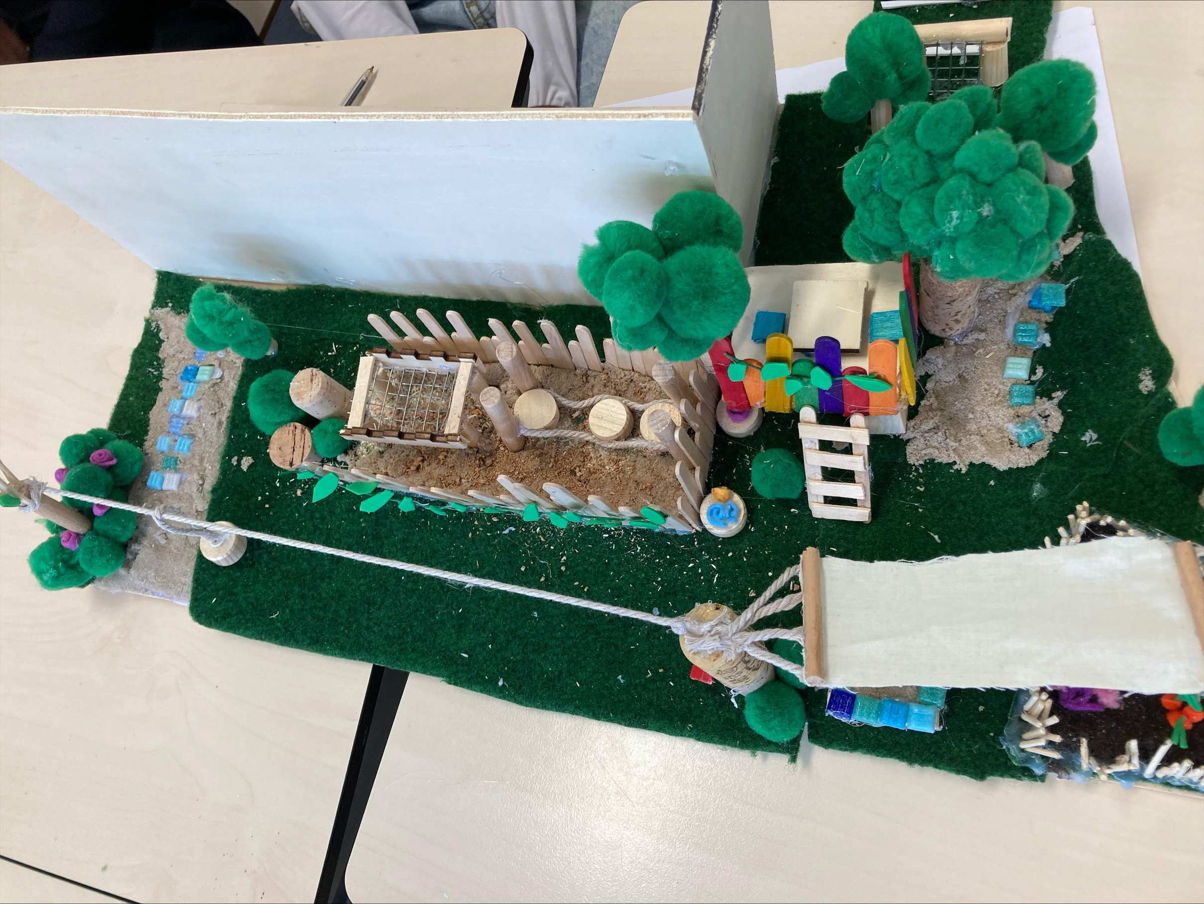 Miniatuurdiorama met tuin, hek, brug en kleurrijke blokken op groen gras.