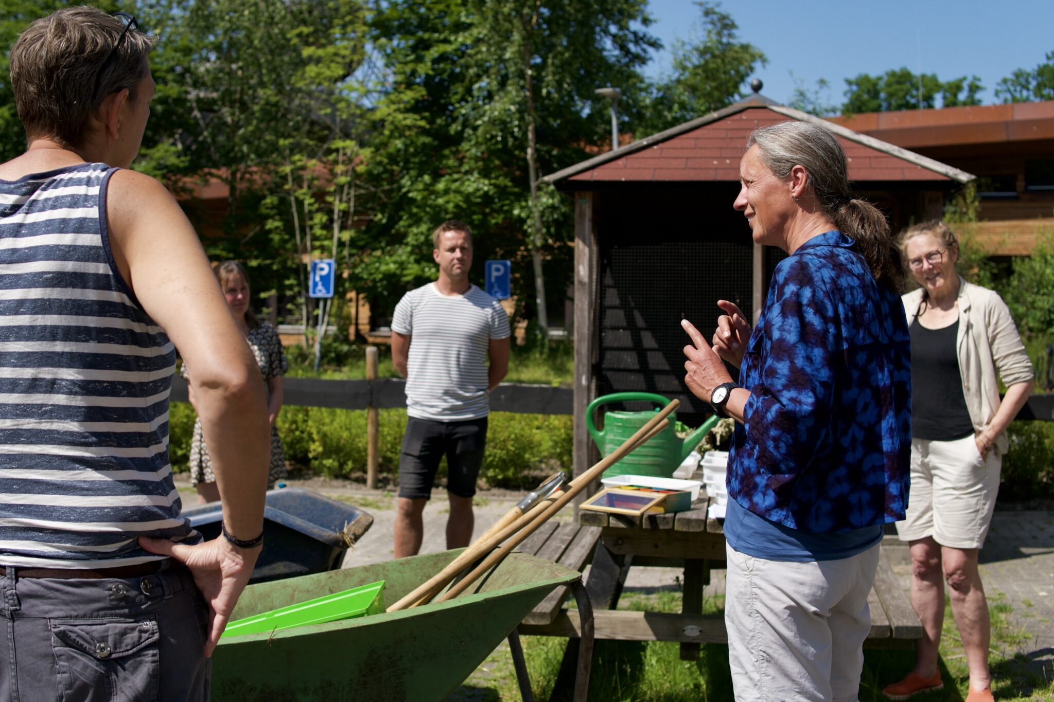 Groep mensen buiten in gesprek, bij een kruiwagen met gereedschap en rolstoelparkeerborden op de achtergrond.
