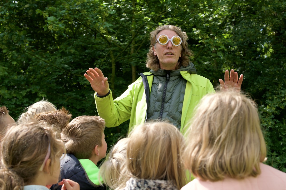 Persoon in felgroene jas spreekt buiten met een groep kinderen, draagt opvallende zonnebril.