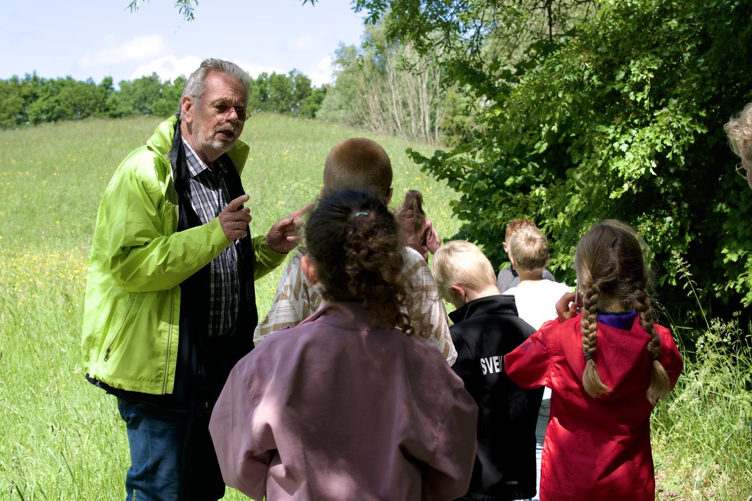 Man in groene jas praat met kinderen in een bosrijke omgeving.