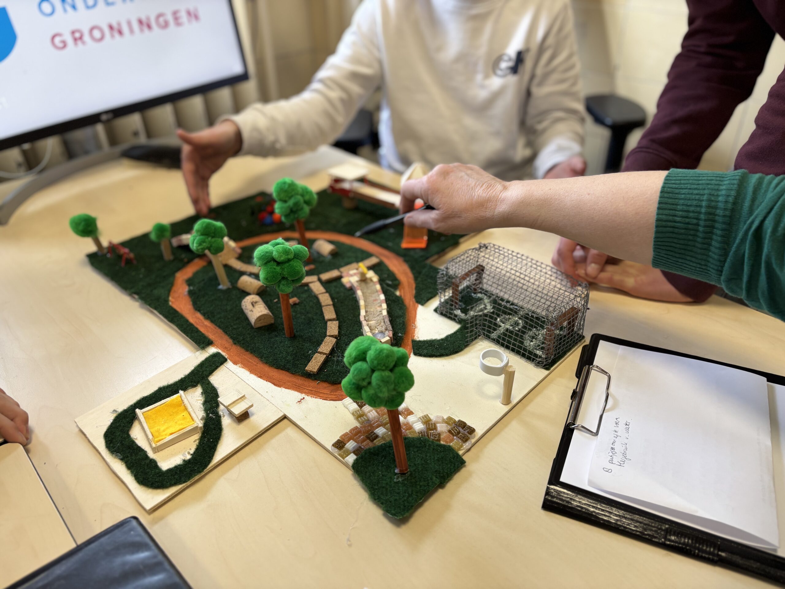 Miniatuurlandschap met bomen en paden; mensen wijzen en bespreken het ontwerp op een tafel.