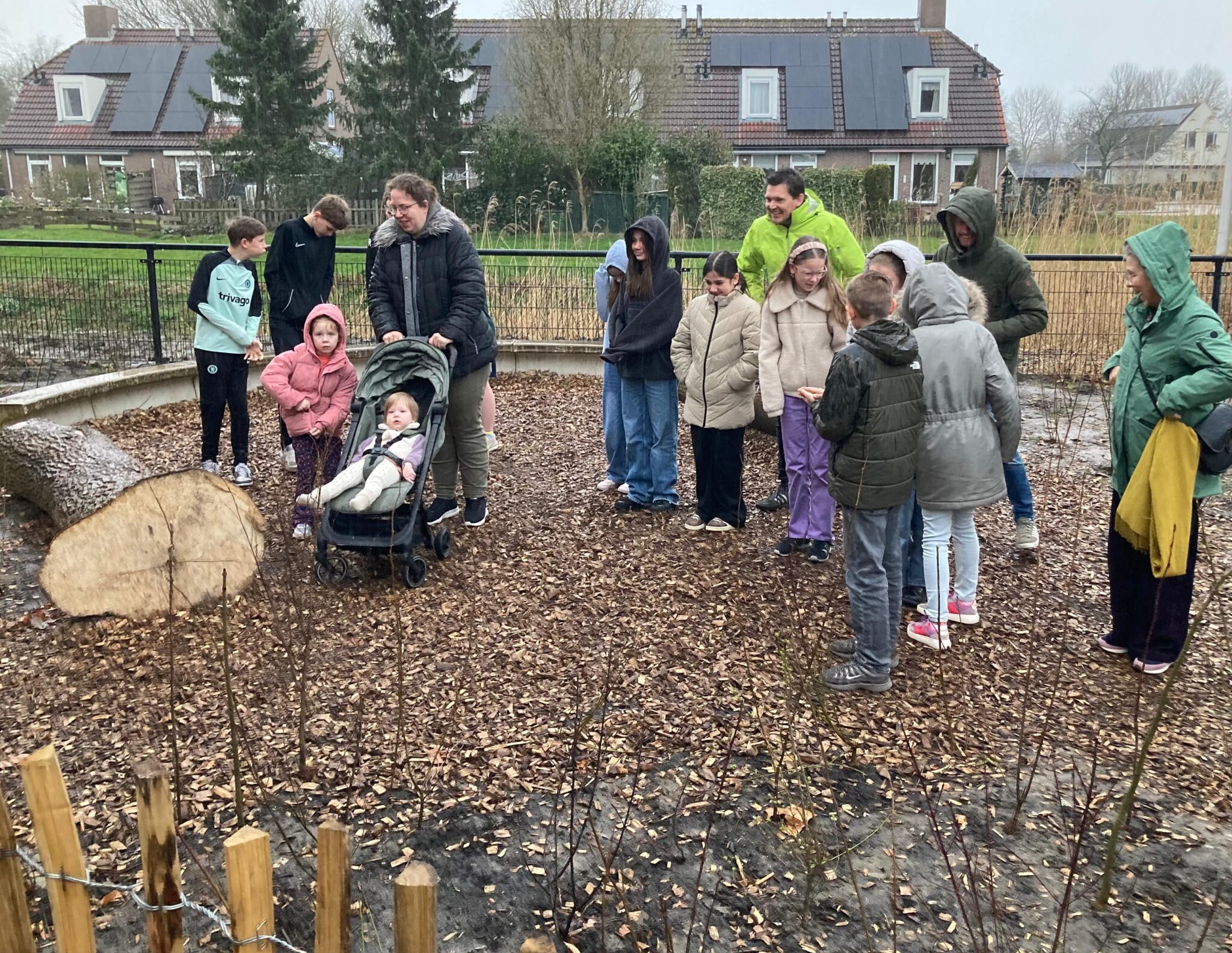 Groep mensen in een tuin, met een kinderwagen, bij een omgevallen boomstam onder een bewolkte hemel.