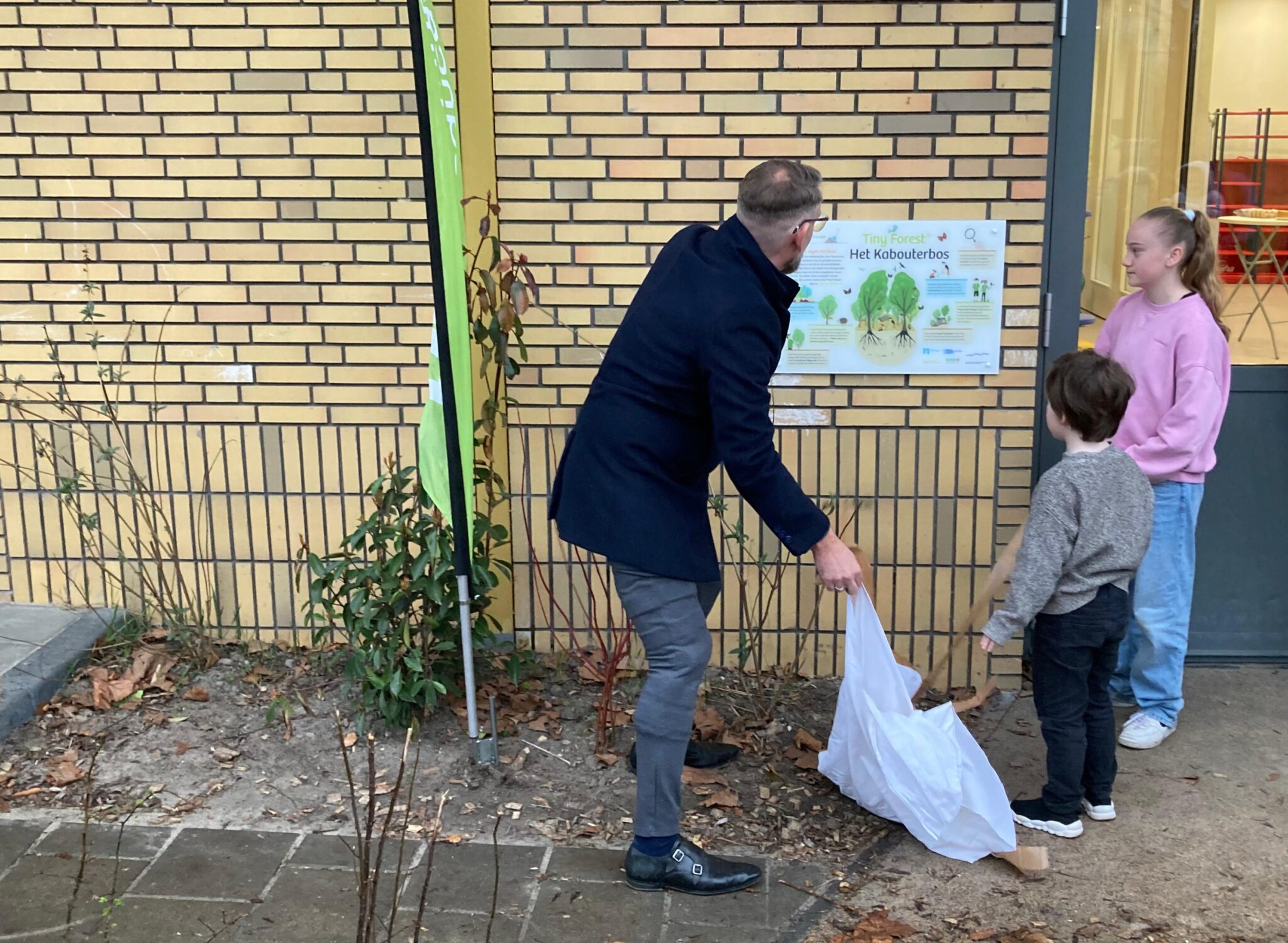 Een man onthult een bord over het Kabouterbos bij een schoolmuur, met twee kinderen naast hem.