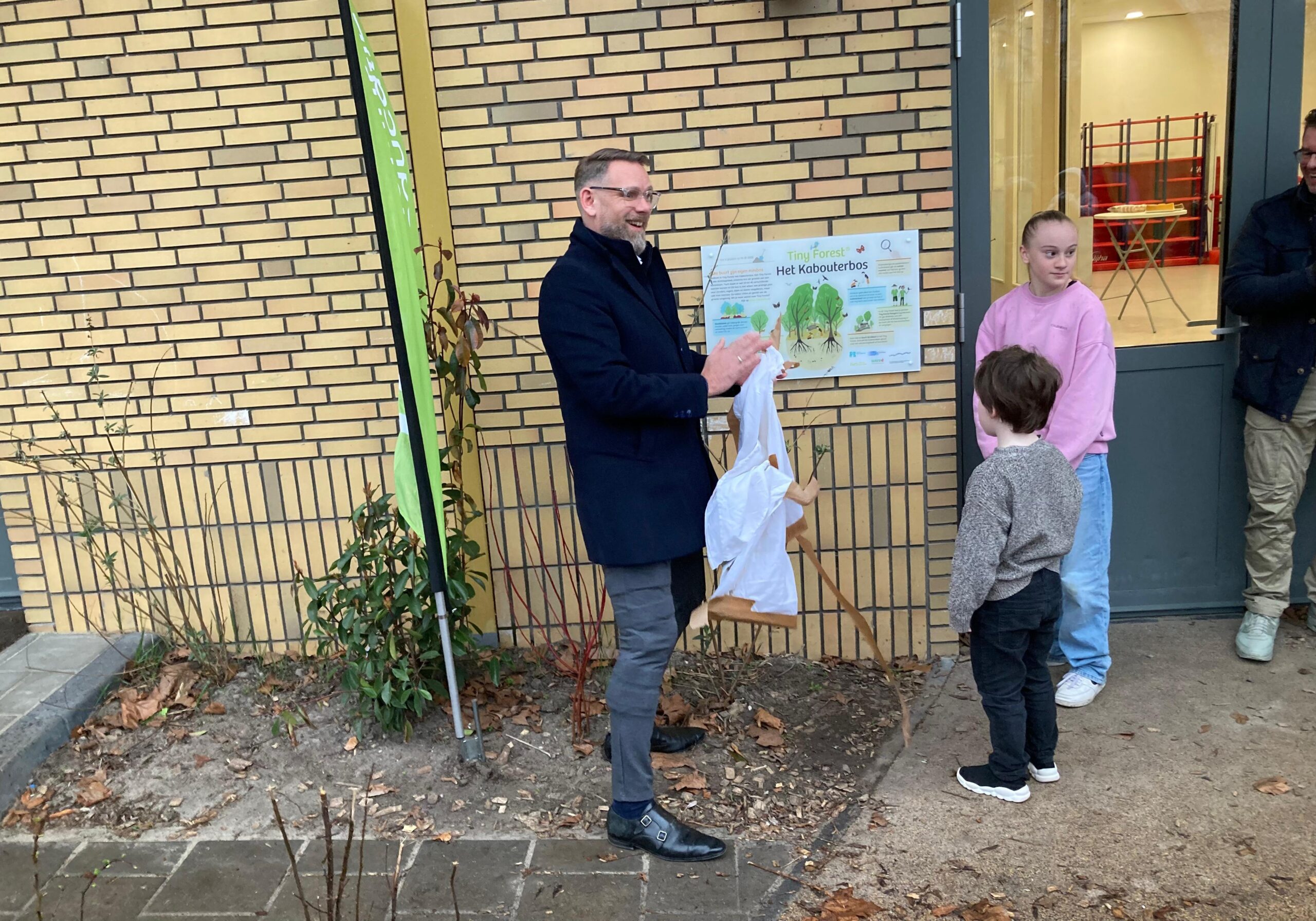 Een man onthult een bord met "Tiny Forest: Het Kabouterbos". Twee kinderen kijken toe.