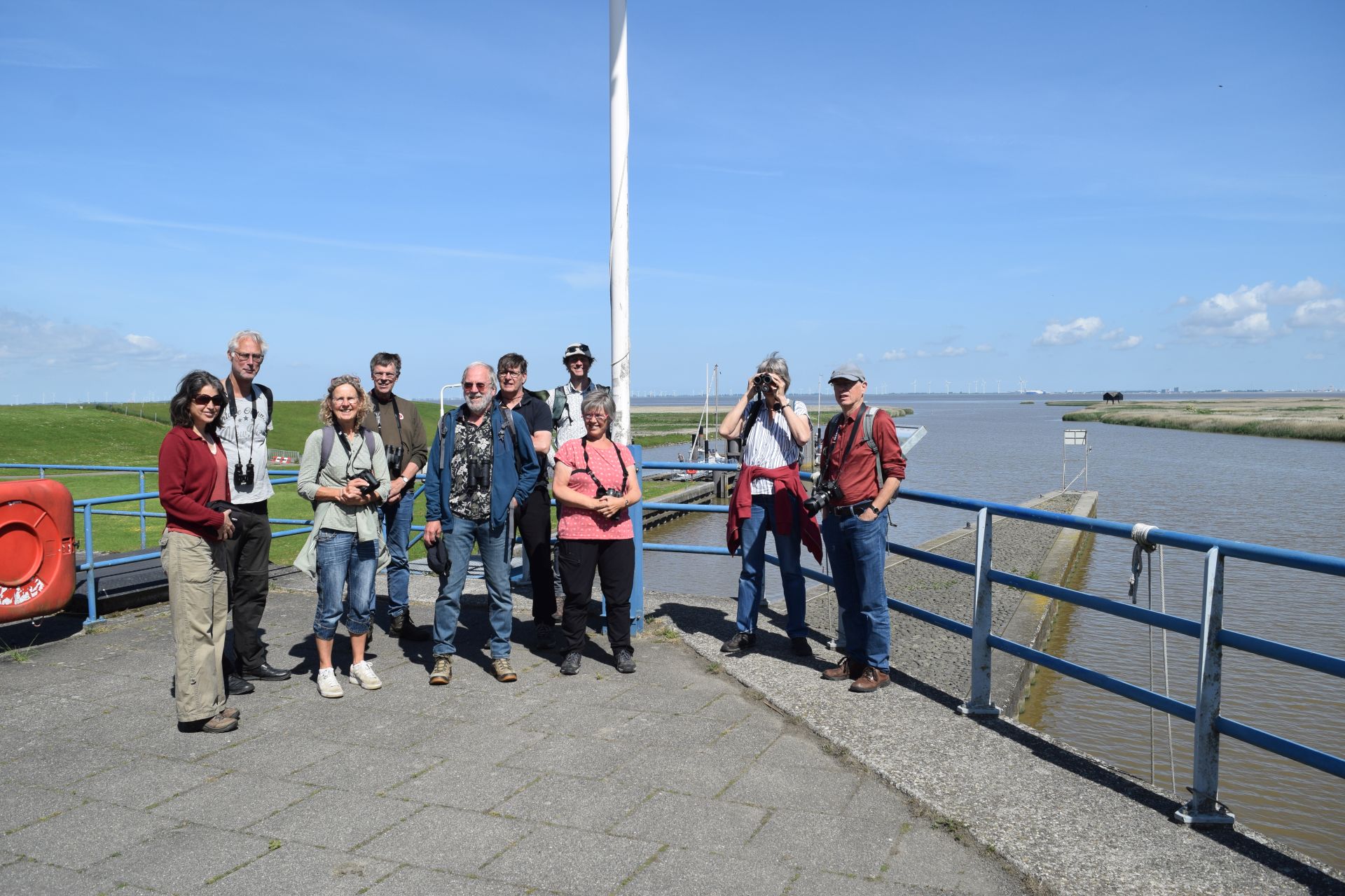Groep van negen mensen met camera's bij een waterkant, met blauwe lucht en wandelpad zichtbaar.