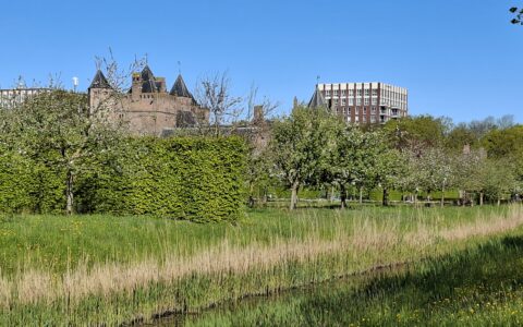 Landschap met kasteel, bomen en modern gebouw onder blauwe hemel.