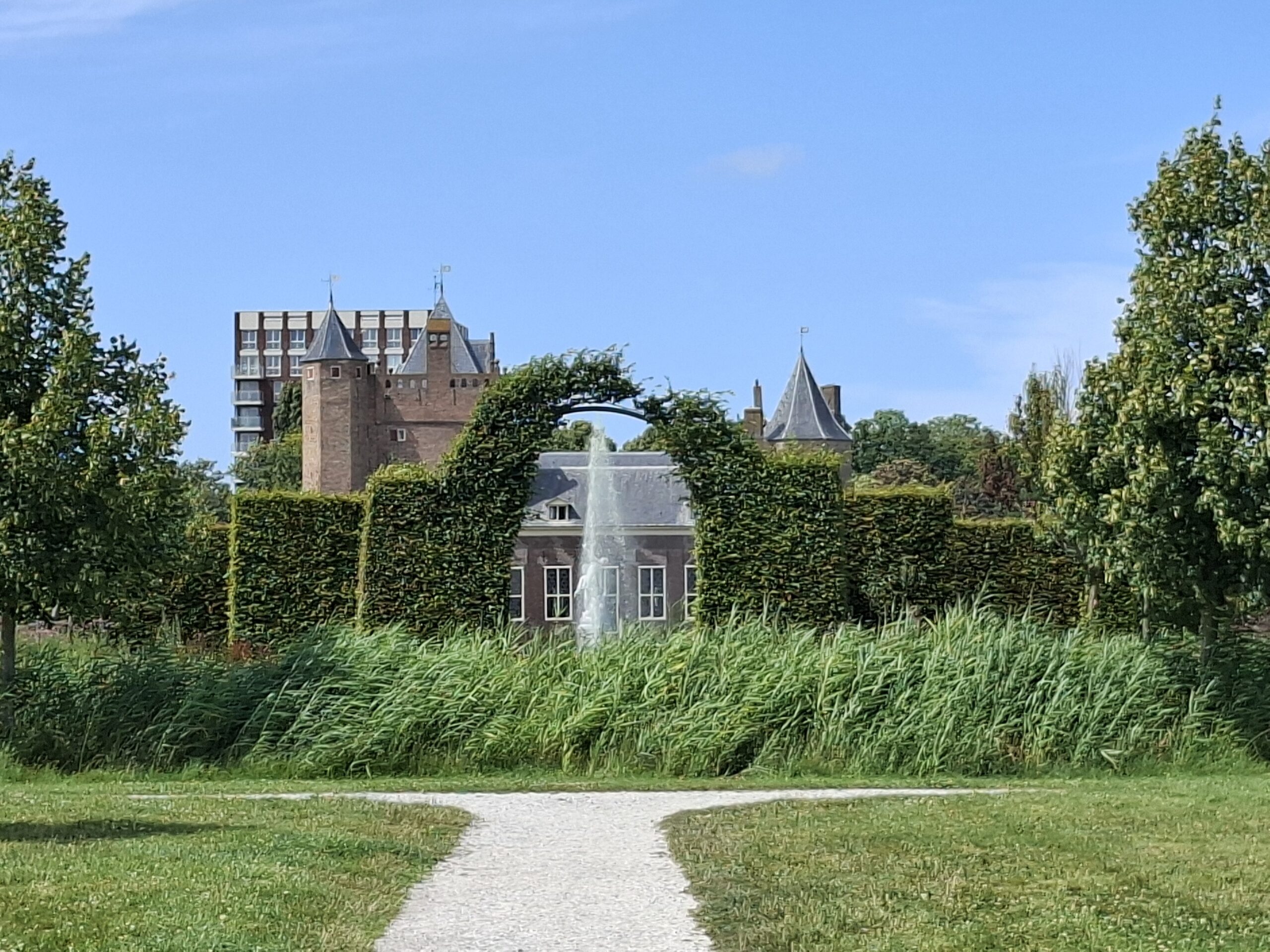 Kasteel met torens achter groene heg en pad, omgeven door bomen, onder blauwe lucht.