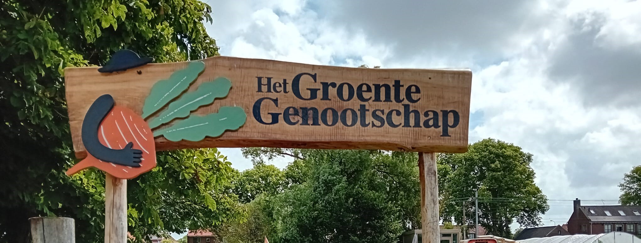 Houten bord met tekst "Het Groente Genootschap" bij een groene, boomrijke achtergrond.
