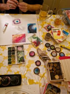 Knutseltafel met aquarelverf, kunstwerken en glazen thee op een gele bloemen tafelkleed.