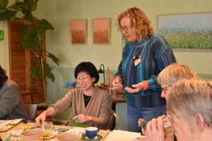 Vrouw in blauwe trui helpt lachende vrouw bij creatieve workshop met kunstbenodigdheden op tafel.