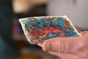 Hand houdt kleurrijk, abstract kunststukje vast; blauw, rood, groen vlekken op gescheurd papier.