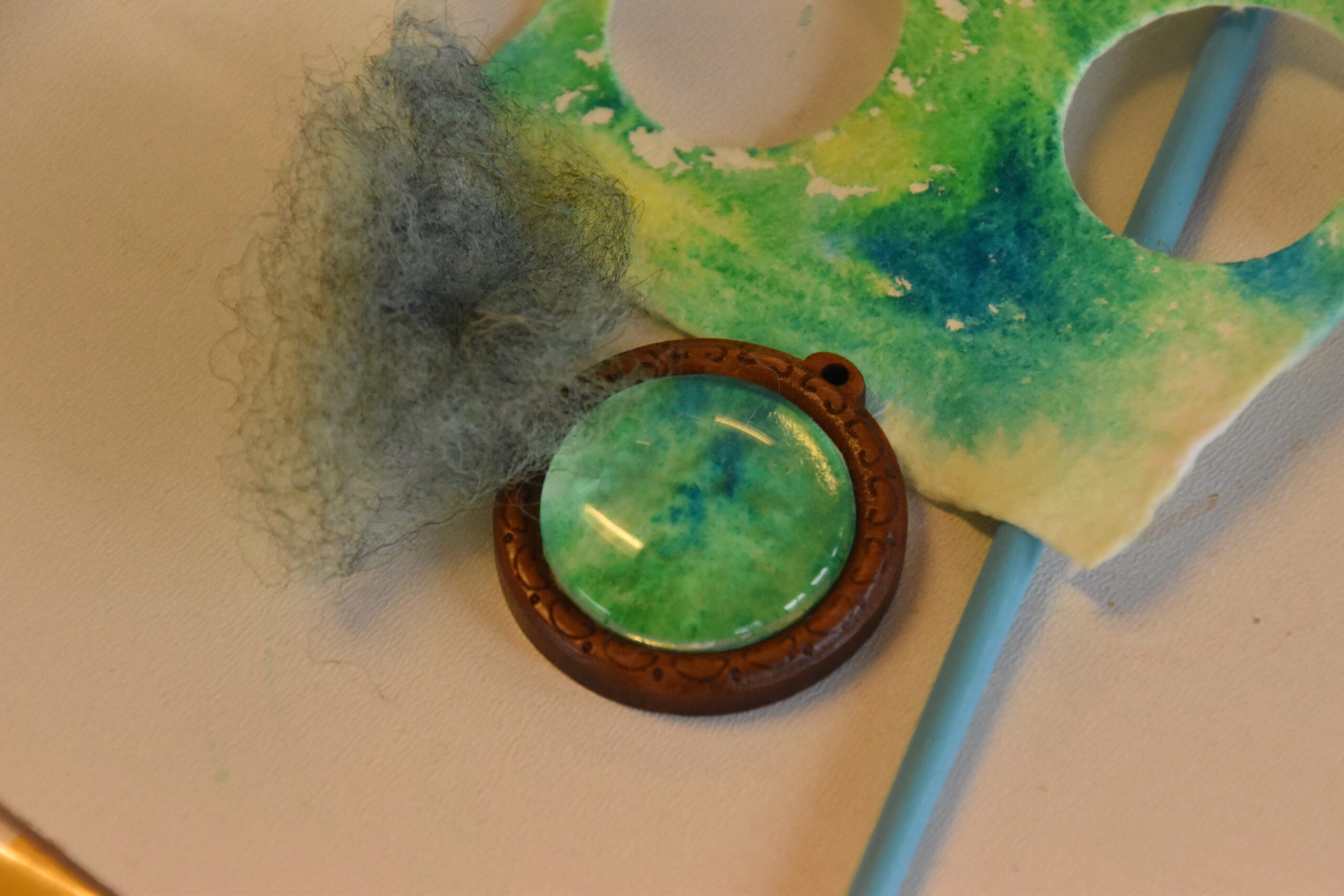 Groene hanger, verfrest op papier, blauwe draad, en stuk grijs vilt op een tafel.