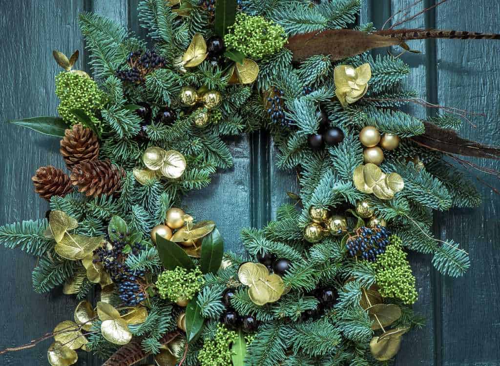 Kerstkrans met dennenappels, bladeren, bessen en gouden decoraties op groene houten deur.