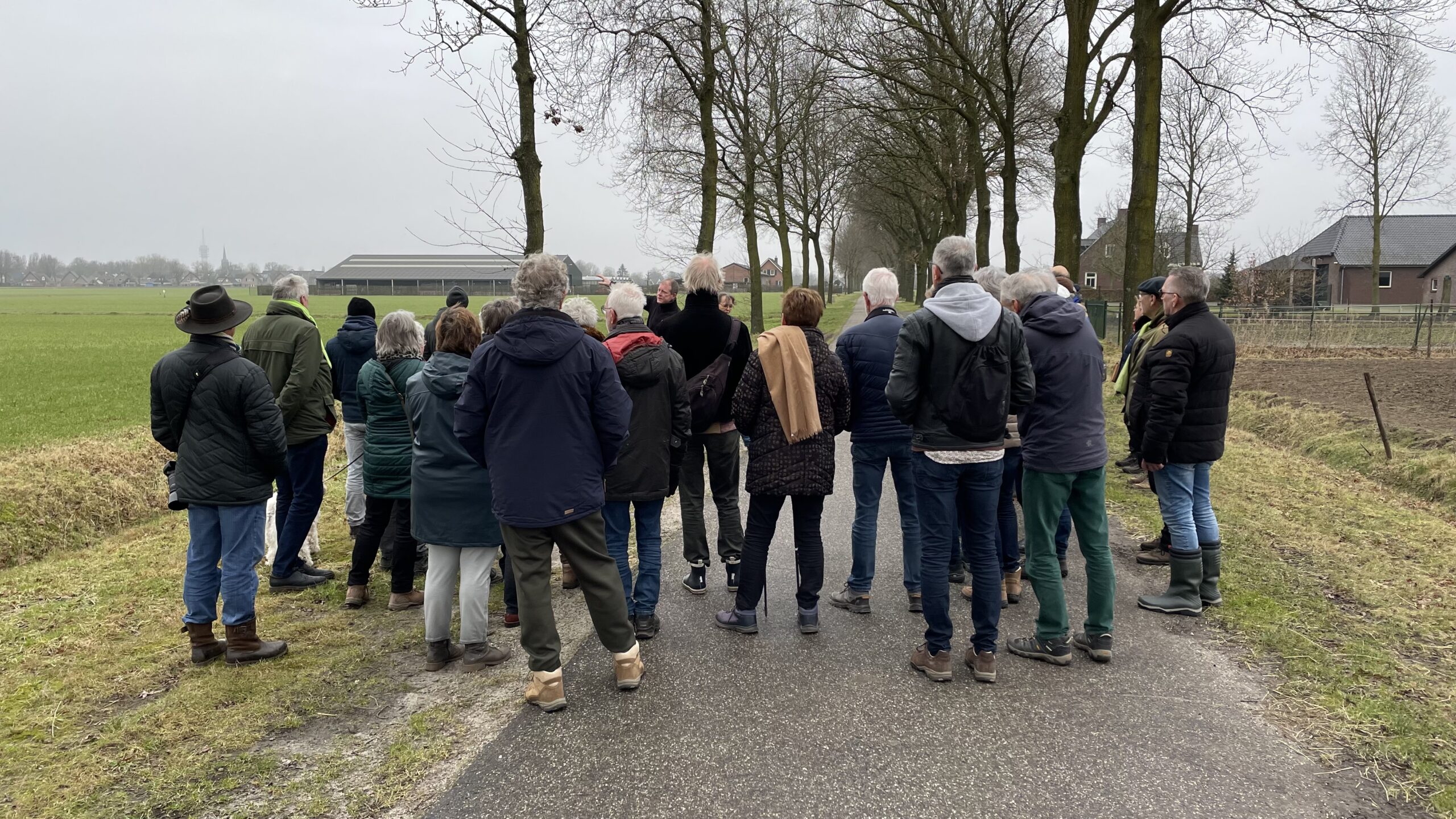 Groep mensen staat op een landweg omgeven door velden en bomen, gericht naar een boerderij.