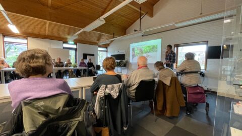 Een groep ouderen volgt een presentatie in een lichte vergaderruimte.