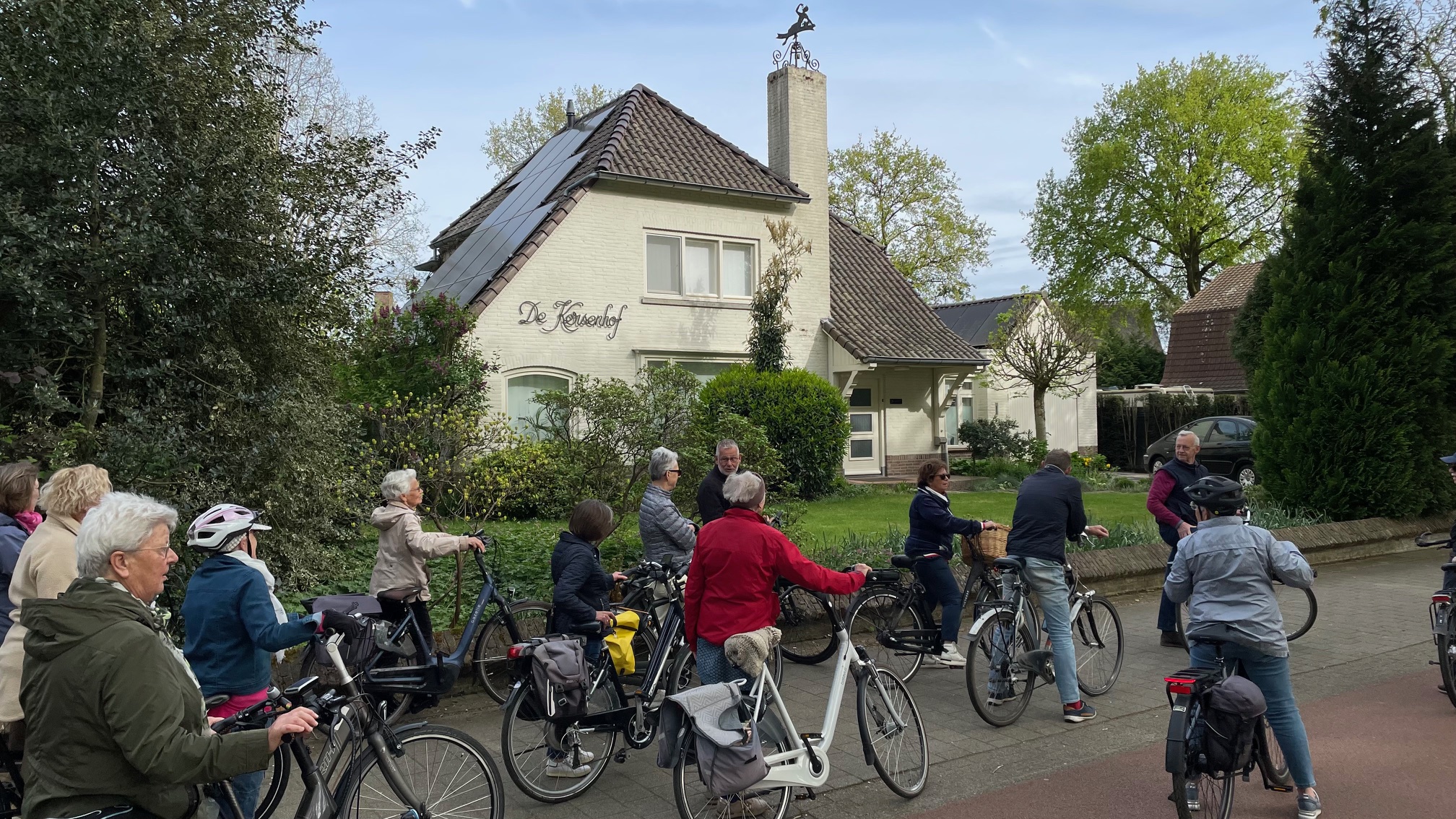 Groep fietsers voor een huis genaamd "De Kersenhof" op een zonnige dag met groene bomen.